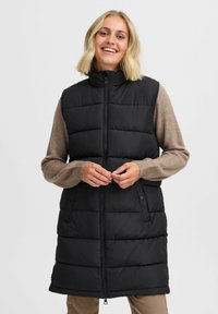 Gilet noir rembourré avec col haut, fermeture éclair et poches latérales. Les manches sont en matière tricotée beige. Texture lisse avec un design structuré.
