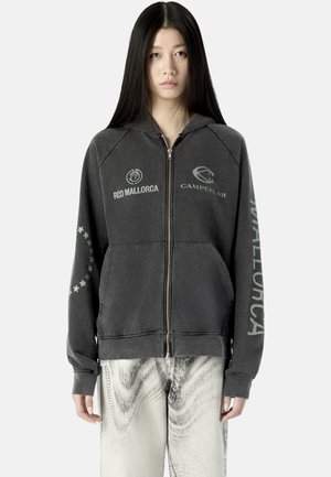 Junge Frau mit langen schwarzen Haaren, die einen dunkelgrauen Kapuzenpullover mit Reißverschluss mit "RCD Mallorca," "Camperlab," und Sternenmuster trägt, kombiniert mit hell gemusterten Hosen.