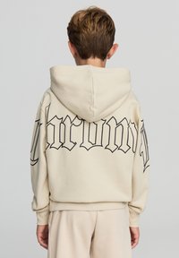 Beżowa bluza z dużą czarną grafiką "Chrono" na plecach, z kapturem na sznurku i ściągaczami na rękawach. Miękki, teksturowany materiał.