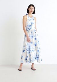 Floralna maksi haljina od bijelog materijala s plavim cvjetnim uzorkom, dizajn s halter vratom i lepršavim siluetom, uparena sa srebrnim sandalama.