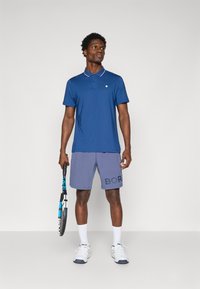 Mornarska polo majica s bijelim ovratnikom, uparena sa sivim kratkim hlačama na kojima je u podebljanom fontu otisnuto "BOR". Model drži tenisku reketa.