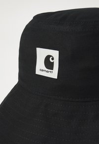 Black bucket hat made of textured fabric featuring a prominent white square patch with a black logo, displaying stitching details and a wide brim.