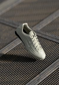 Gaiša pelēka sporta apavi ar struktūru materiālu, saistāmu dizainu un melnu gumijas zoli. Malā ir izteikta swoosh logo.