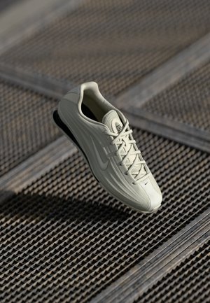Sapatilha atlética cinza claro com parte superior texturizada, design com atacadores e sola de borracha preta. Apresenta um logótipo swoosh proeminente na lateral.