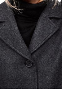Donna che indossa un cappotto di lana grigio scuro con un ampio colletto e un singolo bottone nero su un maglione collo alto nero.