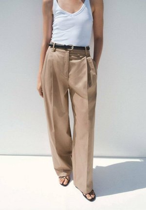 Persona con pantalones beige de pierna ancha, top blanco sin mangas, cinturón negro y sandalias negras con los dedos descubiertos, de pie frente a un fondo liso y claro.