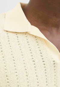 Polo shirt lavorato a maglia color crema con un motivo testurizzato, caratterizzato da un colletto morbido e dettagli di cucitura aperta visibili, visto da un'angolazione ravvicinata.
