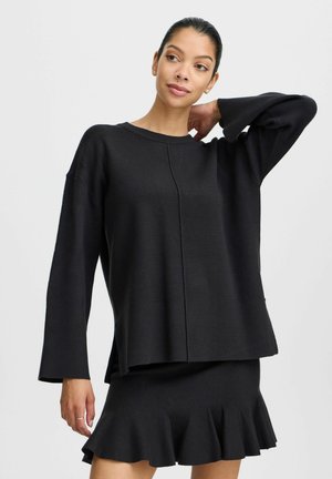 Schwarzer Strickpullover mit überschnittenen Schultern, langen Ärmeln und geradem Saum. Kombiniert mit einem passenden schwarzen Minirock mit weit auslaufendem Saum.