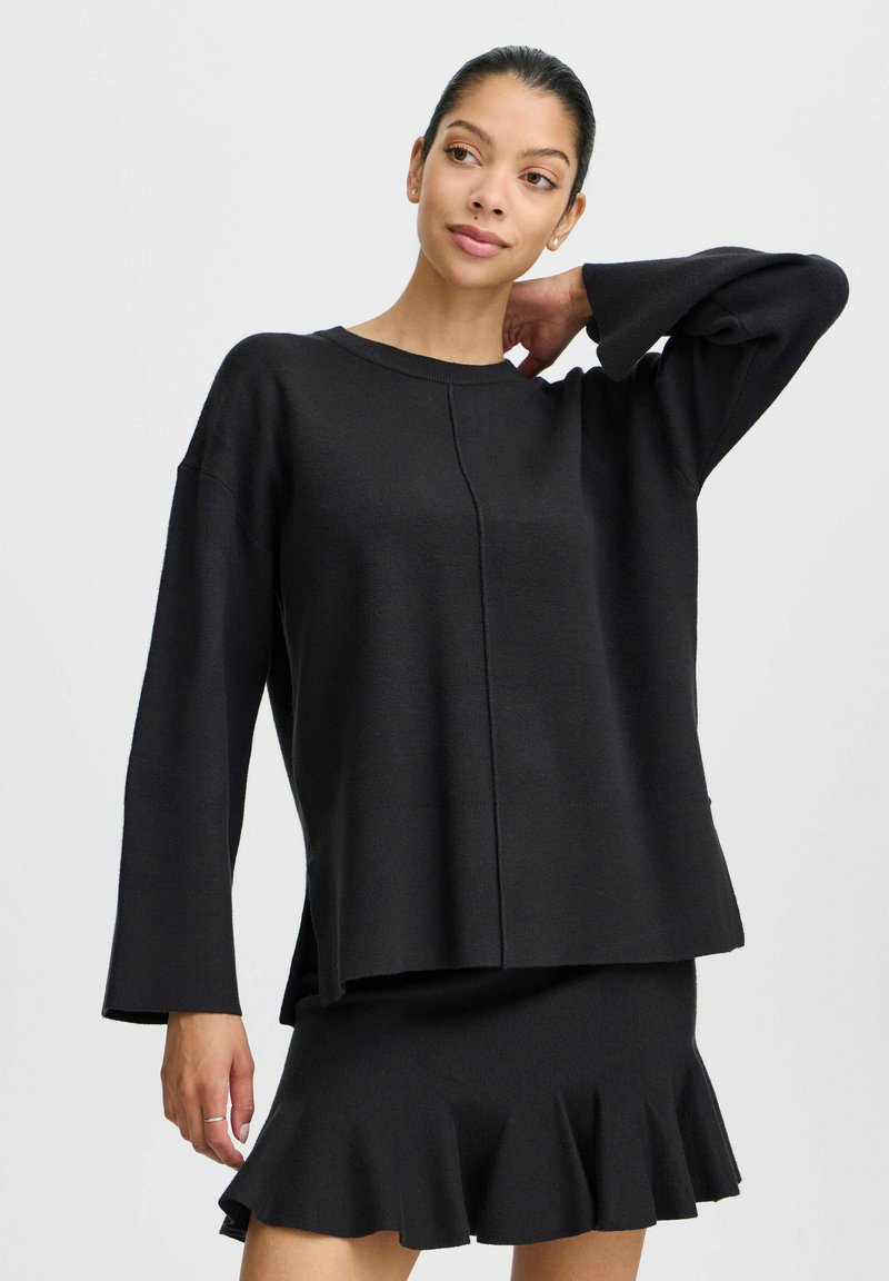 Schwarzer Strickpullover mit überschnittenen Schultern, langen Ärmeln und geradem Saum. Kombiniert mit einem passenden schwarzen Minirock mit weit auslaufendem Saum.