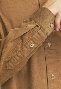 Chemise en velours côtelé marron avec un poignet à bouton, présentant une texture côtelée verticale et un col classique. Comprend deux boutons sur la manche.