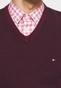 Bordeaux rood gebreide trui met een V-hals ontwerp, gecombineerd met een rood-wit geruit overhemd. Voorzien van een klein logo linksonder op de trui.