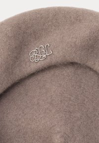 Gaišbrūns filca berets ar gludu tekstūru. Malā ir pievienots sudraba monograms "PBL". Apaļa forma.