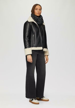 Schwarze Lederjacke mit crèmefarbigem Lammfellkragen und -manschetten, schwarzer Schal, schwarze weit geschnittene Hosen und beige Sneakers mit schwarzen Akzenten.