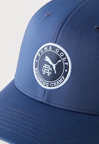 Μπλε καπέλο γκολφ με κεντημένο λογότυπο Puma και κείμενο "Puma Golf Reigning Champ" σε κυκλικό πατς στο μπροστινό πάνελ.