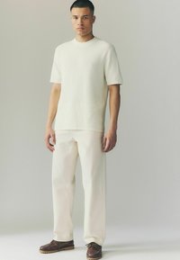 Short-sleeve cream knit shirt paired with white trousers and brown leather moccasin shoes. Simple, modern design with minimal detailing.