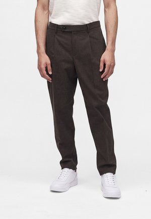 Mand iført mørkebrune skræddersyede bukser og hvide sneakers, stående mod en ensfarvet hvid baggrund.