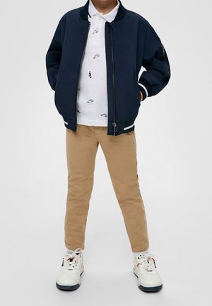 Veste bomber bleu marine, polo blanc avec motifs de voitures, pantalon beige et baskets blanches avec des accents rouges et bleus. Lignes épurées et design décontracté.