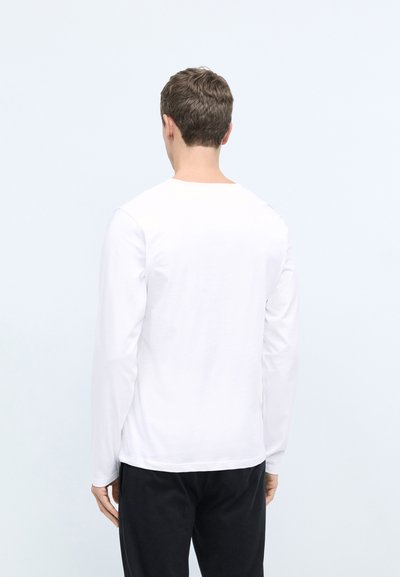 T-shirt blanc à manches longues en coton, avec un col rond et un ourlet droit, porté avec un pantalon sombre, montrant la vue arrière.