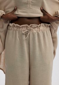 Conjunto de lino beige con una cintura fruncida y cordón. La parte superior tiene un cierre de botón y mangas fruncidas. Textura suave.