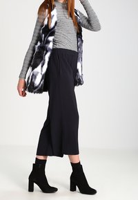 Czarny i biały kamizelka z futra syntetycznego noszona na paskowanej bluzce z długim rękawem, w połączeniu z czarnymi spodniami o szerokich nogawkach i czarnymi botkami za kostkę.