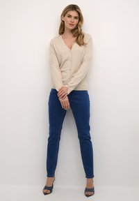 Beige cardigan with v-neck and button front, paired with blue fitted jeans. The model wears navy open-toe heels, showcasing a smooth texture.