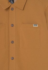 Brązowa polarowa koszula z kołnierzykiem i dwoma przednimi kieszeniami, z ciemnymi guzikami i małą metką "Converse All-Star" na lewej kieszeni.