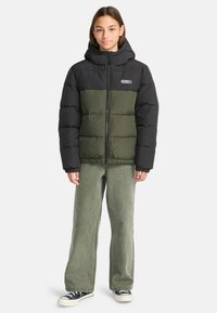 Giacca a vento nera e verde oliva con cappuccio, caratterizzata da un design trapuntato, zip frontale e patch con logo. Indossata sopra pantaloni larghi verde chiaro.