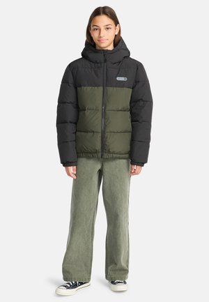 Zwarte en olijfgroene pufferjack met capuchon, voorzien van een doorgestikte ontwerp, vooraan een rits en een logo afbeelding. Draagt over lichtgroene wijde broek.