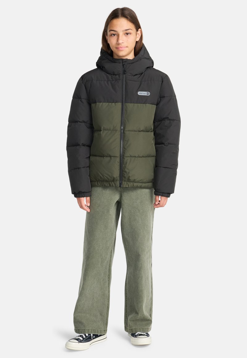 Giacca a vento nera e verde oliva con cappuccio, caratterizzata da un design trapuntato, zip frontale e patch con logo. Indossata sopra pantaloni larghi verde chiaro.