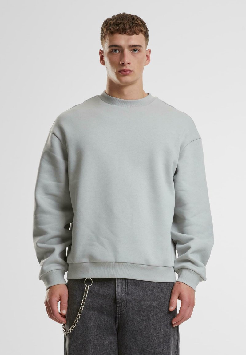 Hellgrauer, übergroßer Pullover mit rundem Kragen, gerippten Bündchen und Saum. Trägt dazu eine dunkelgraue Hose mit Kettendetail.