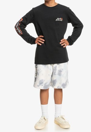 Dziecko ubrane w czarną koszulkę z długim rękawem Quiksilver z logo, szare i białe krótkie spodenki tie-dye z logo, białe skarpetki oraz beżowe trampki, stojące z rękami na biodrach.