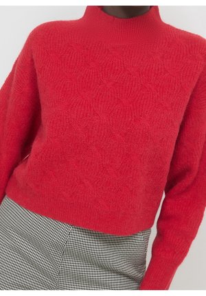 Pull en maille texturée rouge à manches longues associé à une jupe à motif pied-de-poule noir et blanc.