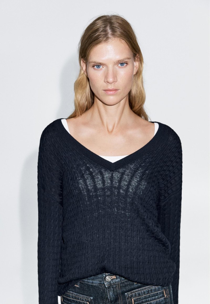 Blonde Frau mit blauen Augen, die einen schwarzen, strukturierten V-Ausschnitt-Pullover über einem weißen Oberteil und dunklen Jeans trägt, vor einem schlichten weißen Hintergrund stehend.