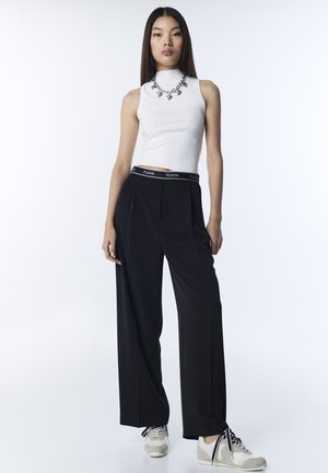 Pantalon noir à jambes larges avec une ceinture brandée FLEUR, associé à un haut blanc sans manches et des baskets grises. Mis en valeur par un collier argenté.