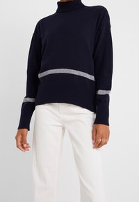 Person in einem marineblauen Pullover mit grauen Streifen an den Ärmeln und am Saum, kombiniert mit weißen Hosen vor einem einfarbigen hellen Hintergrund.