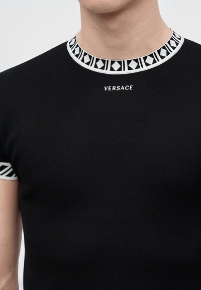 Κοντινό πλάνο ενός μαύρου t-shirt Versace με γεωμετρικό ασπρόμαυρο σχέδιο στις άκρες του γιακά και των μανικιών.