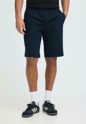 Mężczyzna ubrany w granatowe krótkie spodenki do kolan, białe skarpetki do kostek oraz czarne buty Adidas z białymi paskami, stoi z rękami w kieszeniach.