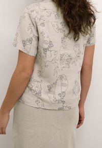 Hedvábná košile s krátkým rukávem v jemné béžové barvě s nepretržitým vzorem kontur obličejů a rostlin. Spodní lem je zakřivený a střih je volný.