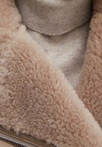 Beżowy kołnierz z barankiem o miękkiej, puszystej fakturze, nałożony na jasnobeżowy sweter z golfem. Na kurtce widoczny jest detal z zamkiem błyskawicznym.