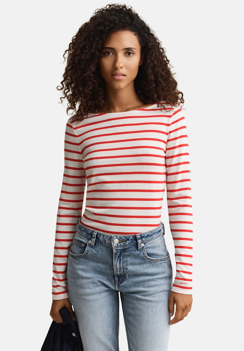 Γυναίκα με μπούκλες φοράει λευκό μπλουζάκι με κόκκινες οριζόντιες ρίγες και ανοιχτό μπλε τζιν, κρατώντας ένα σκουρόχρωμο ύφασμα στο αριστερό της χέρι.