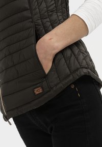 Donkerbruin gewatteerd vest met een geborduurd patroon, zijzakken en een klein leren logo-label nabij de rits. Zwarte broek zichtbaar aan de onderkant.
