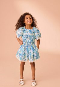 Blommig klänning i blå och gröna nyanser, med puffärmar, smockad midja och spetsdetaljer i fållen. Matchad med vita sandaler.