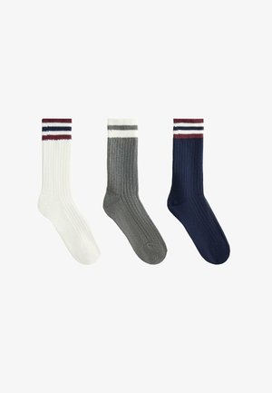 Tri para rebrastih čarapa: jedan bijeli s crvenim i tamnoplavim prugama, jedan sivi i jedan tamnoplavi. Mekana, pletena tekstura, klasična visina do gležnja.