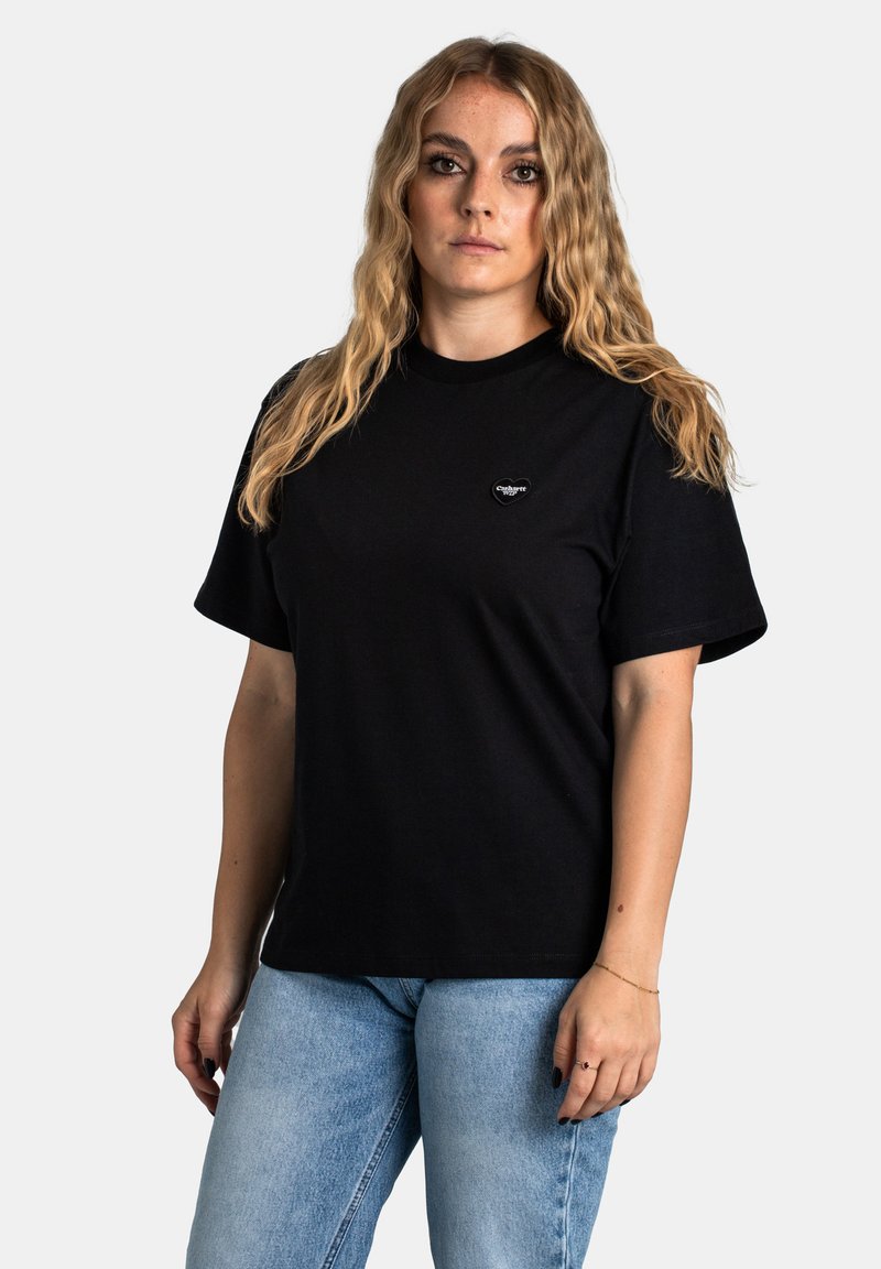 Schwarzes Baumwoll-T-Shirt mit kurzen Ärmeln, Rundhalsausschnitt und einem kleinen Herz-Logo auf der Brust. Kombiniert mit hellblauen Jeans.