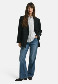 Donna che indossa un blazer nero, una camicia bianca con bottoni, jeans larghi blu e scarpe nere a punta in piedi contro uno sfondo bianco.