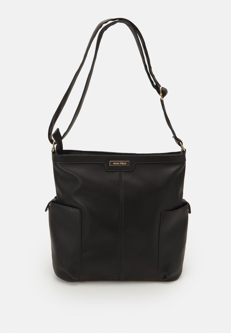 Sac à main en cuir noir avec une bandoulière réglable, deux poches latérales et une petite étiquette dorée indiquant « Anna Field » à l'avant.