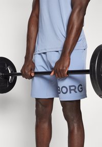 Calções de desporto azul claro com o logótipo "BORG", barra de peso preta segurada com duas mãos, destacando braços musculosos e pele texturizada.