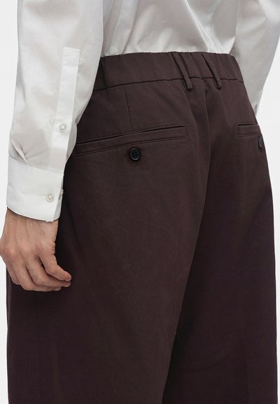 Gros plan d'une personne portant un pantalon marron foncé avec des poches arrière boutonnées et une chemise blanche à manches longues rentrée.