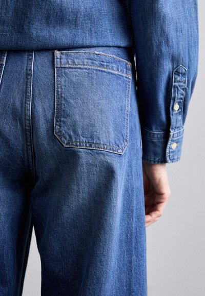 Blue denim jeans with a classic design, featuring a back pocket with top stitching, and a smooth texture; side visible with a fitted silhouette.