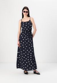 Rochie maxi din material bleumarin cu buline albe, cu bretele spaghetti ajustabile și talie elastică. Asortată cu pantofi joși negri și ochelari de soare.